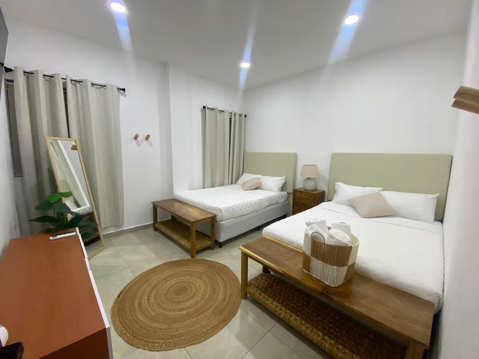 A hotel room with two beds, a desk, a mirror, a plant, and a woven rug.