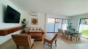 Ein modernes Wohnzimmer mit einer Couch, zwei Korbstühlen, einem Couchtisch, einem an der Wand montierten Fernseher und einem Essbereich mit Tisch und Stühlen.