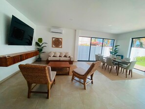 A modern living room with a sofa, two wicker chairs, a coffee table, a TV mounted on the wall, and a dining area with a table and chairs. - Beachfront Luxury Villa with Private Pool in the fantastic Costa Del Sol (San Pedro Masahuat)