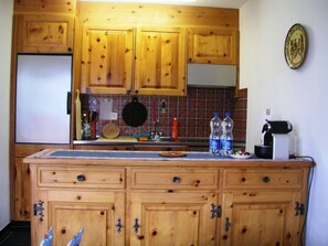 A kitchen with wooden cabinets, a tiled backsplash, a refrigerator, a coffee maker, and two water bottles. - Köppel, (Disentis/Mustér), 1053 by Interhome (Disentis)