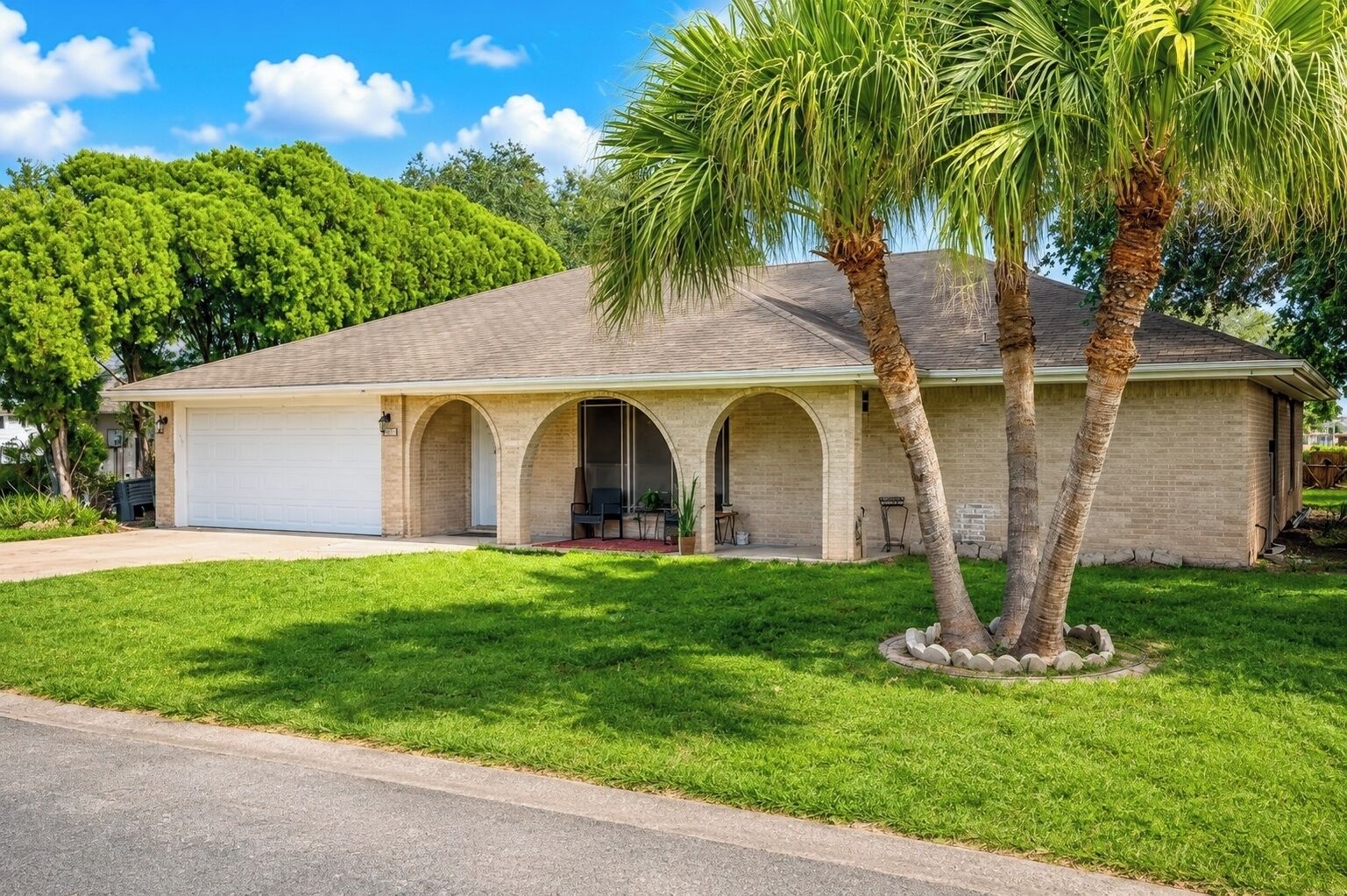 A single-story house with a garage, a covered porch with arches, and a lawn with palm trees.