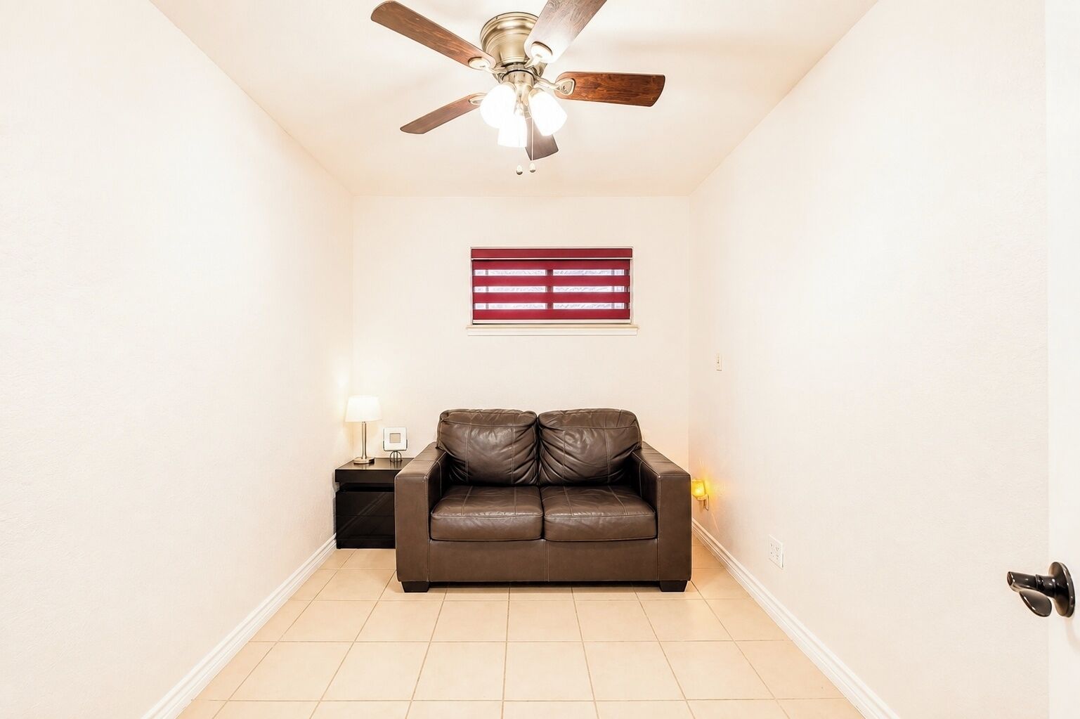 Um quarto com ventilador de teto, janela com persianas, um sofá de couro marrom, uma mesa pequena com abajur e uma luminária na parede.