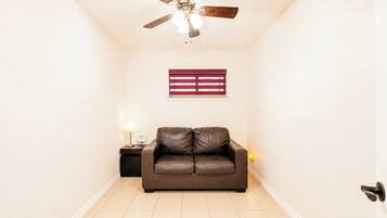 Um quarto com ventilador de teto, janela com persianas, um sofá de couro marrom, uma mesa pequena com abajur e uma luminária na parede.