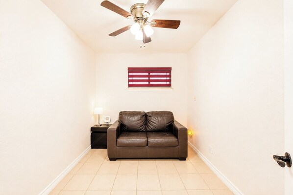 A room with a ceiling fan, a window with blinds, a brown leather sofa, a small table with a lamp, and a wall-mounted light.