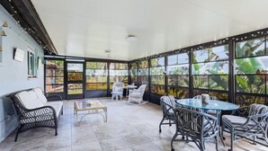 Eine überdachte Terrasse mit Korbmöbeln, einem Glastisch und großen Fenstern mit Blick auf eine tropische Landschaft.