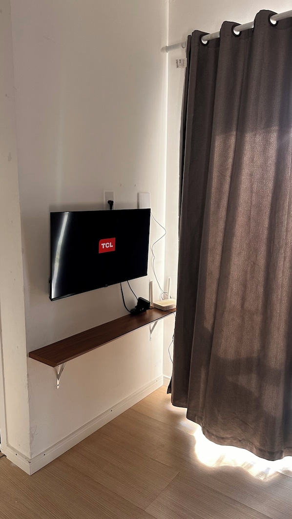 A flat-screen TV mounted on a wall, a power outlet, a cable management system, a wooden shelf, and a window with curtains.