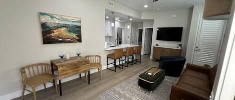 A modern living room with a dining area, a ceiling fan, a flat-screen TV, and a kitchen in the background.