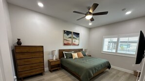 Quarto com cama, penteadeira de madeira, mesas de cabeceira, ventoinha no teto e janela.