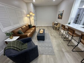 Ein modernes Wohnzimmer mit einem braunen Ledersofa, einem dunklen Ohrensessel, einem Holztisch und einem Essbereich mit Barhocker.