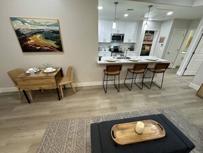 Ein Essbereich mit Holztisch und -stühlen, eine Küche mit weißen Schränken und einer zentralen Kochinsel sowie ein Gemälde an der Wand.