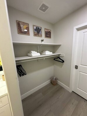Ein kleines, sauberes Badezimmer mit weißen Wänden, Holzboden, einem Handtuchhalter, einem Regal mit gefalteten Handtüchern und einer Tür.