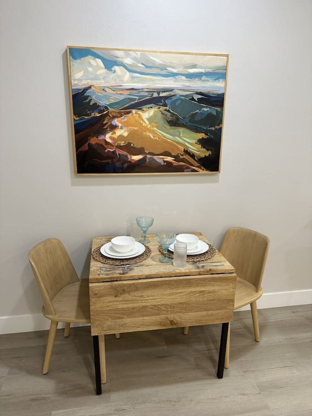 A dining table with two chairs, a painting on the wall, and a wooden floor.