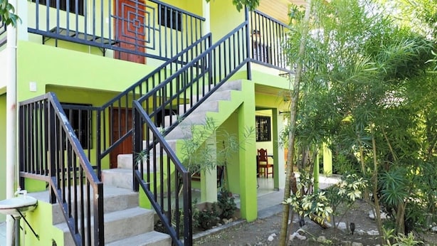 A two-story building with a staircase leading to the upper floor, surrounded by a garden with various plants and a small pond. - Grenn House (Sámara)