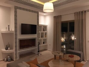 Ein modernes Wohnzimmer mit Flachbildfernseher, Kamin, Essbereich mit Tisch und Stühlen sowie einem großen Fenster mit Vorhängen.
