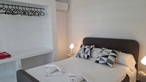 Een net opgemaakt bed met kussens, een nachtkastje met een lamp, een handdoek op het bed, en een kapstok met kleding.