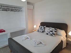 Ein ordentlich bezogenes Bett mit Kissen, ein Nachttisch mit Lampe, ein Handtuch auf dem Bett und ein Kleiderständer mit Kleidung.