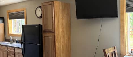 A compact kitchen with wooden cabinets, a black refrigerator, a wall-mounted TV, and a dining area with a table and chairs.