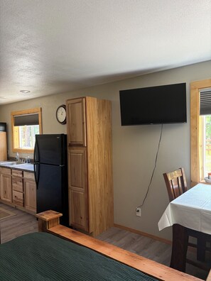 A compact kitchen with wooden cabinets, a black refrigerator, a wall-mounted TV, and a dining area with a table and chairs.