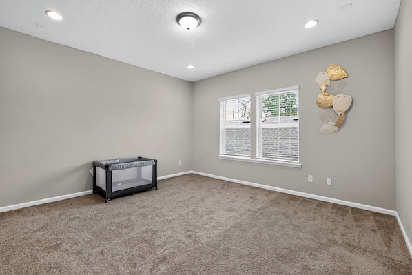 A room with a carpeted floor, a playpen, two windows with blinds, and a ceiling with recessed lighting.
