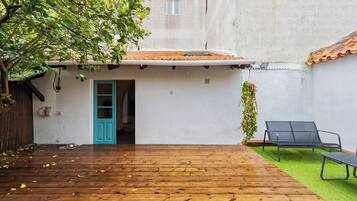 A wooden deck with a blue door leading to a white building, a hanging plant, and a black lounge chair.