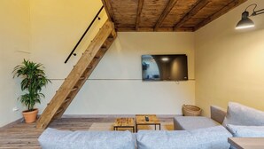 A modern living room with a sloped ceiling, a large flat-screen TV mounted on the wall, a wooden staircase, and a seating area with a sofa and a small table. - Palmela Oasis Apartment II, By Home Match (Palmela)