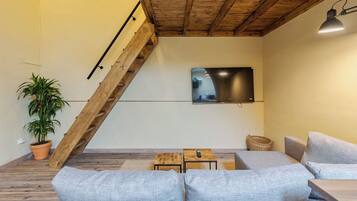 A modern living room with a sloped ceiling, a large flat-screen TV mounted on the wall, a wooden staircase, and a seating area with a sofa and a small table.