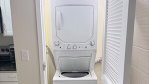 A laundry room with a front-loading washer and dryer, a door leading to a kitchen, and a tiled floor. - Newly Renovated Studio Apartment in Dowtown Roanoke (Roanoke)