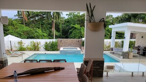 Een buitenzwembad met een houten tafel en stoelen, een jacuzzi en een barbecue.