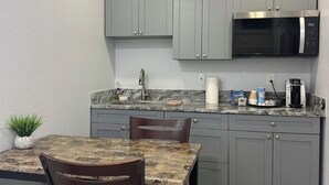 A compact kitchen with grey cabinets, a built-in microwave, a granite countertop, and a dining area with a table and chairs. - Private 1BR Suite, Free Parking, Near Downtown & Airport (Tampa)