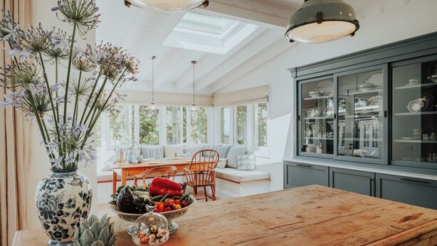 Una cocina con una mesa de madera, un bol de fruta, un florero y un armario con puertas de vidrio.