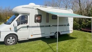 Un camping-car blanc est garé sur l’herbe, avec un espace de vie abrité et une petite fenêtre.