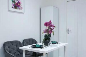 Un comedor con una mesa blanca, dos sillas grises, un jarrón con flores moradas y un armario al fondo.