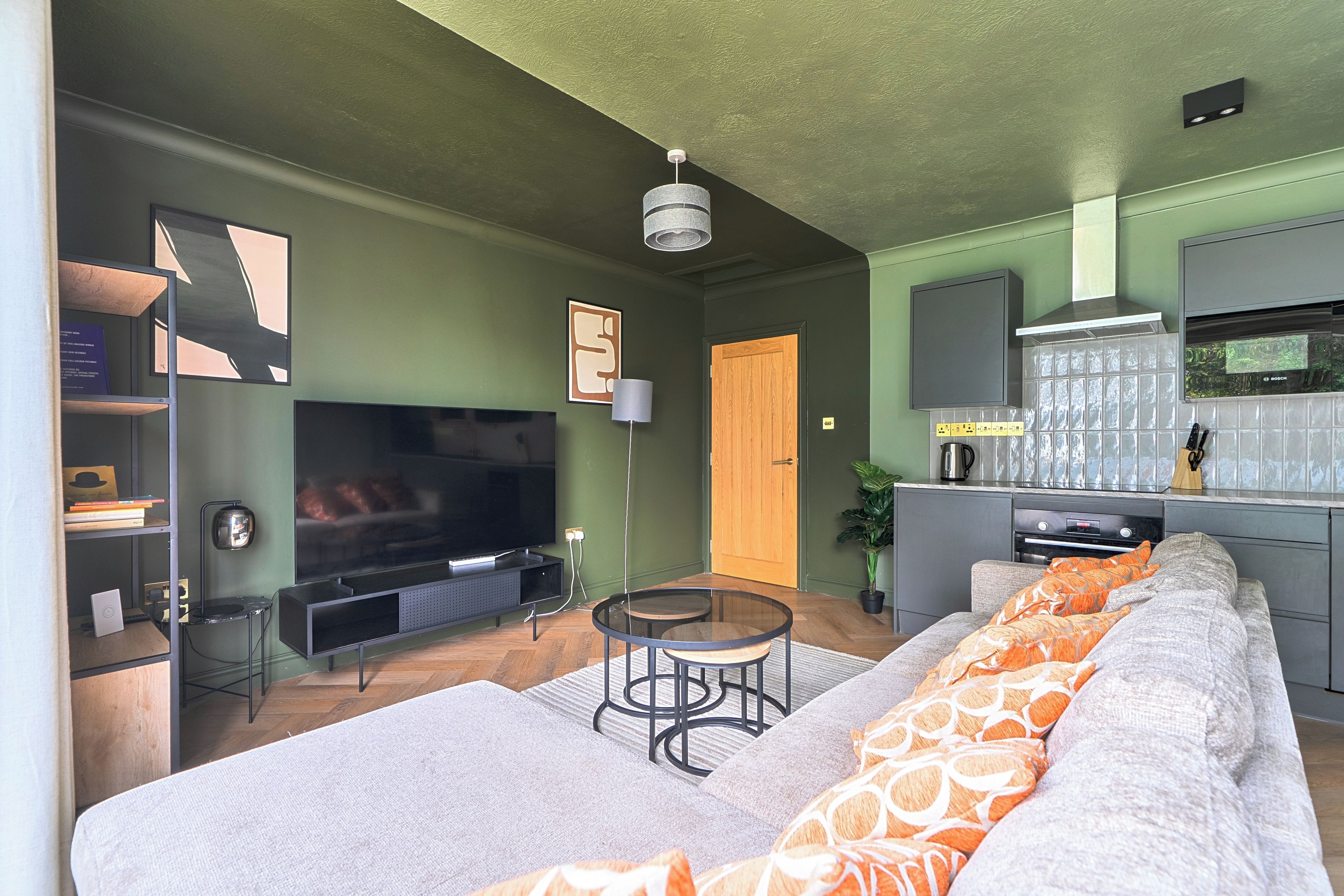 A modern living room with a flat-screen TV, a sofa with patterned cushions, a coffee table, and a kitchen area with a Bosch oven.