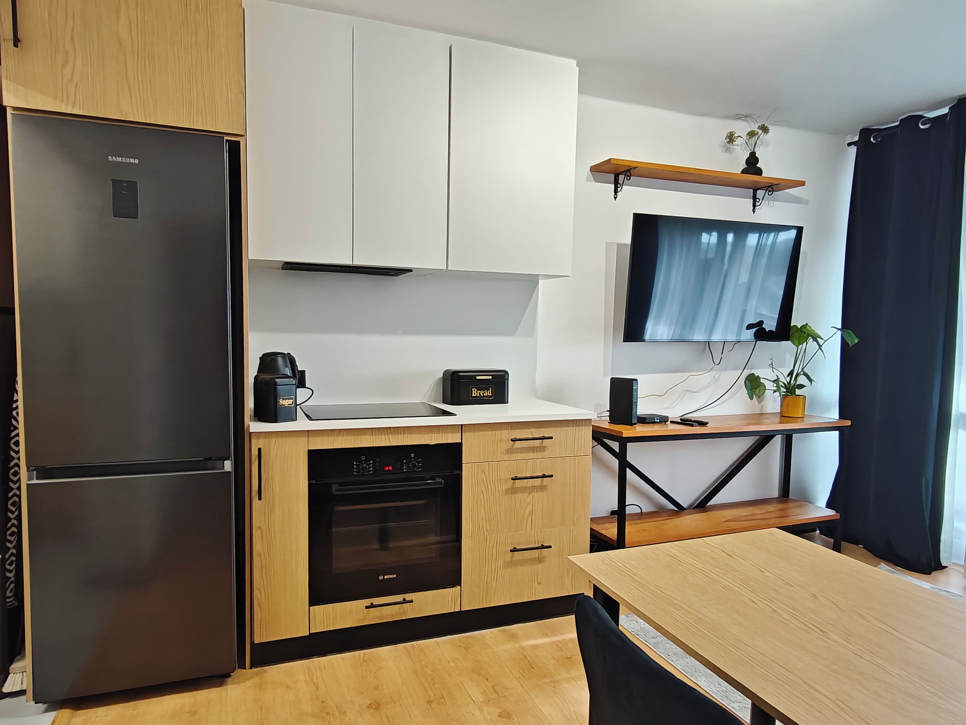 A modern kitchen with a refrigerator, oven, and a coffee maker. There is a wooden dining table and a TV mounted on the wall.