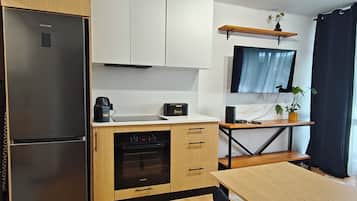 A modern kitchen with a refrigerator, oven, and a coffee maker. There is a wooden dining table and a TV mounted on the wall.