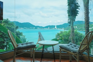 A room with a view of a coastal landscape, including a body of water and distant hills. There are two wicker chairs and a small round table in the foreground. - Blue Harbour Phuket (Patong)