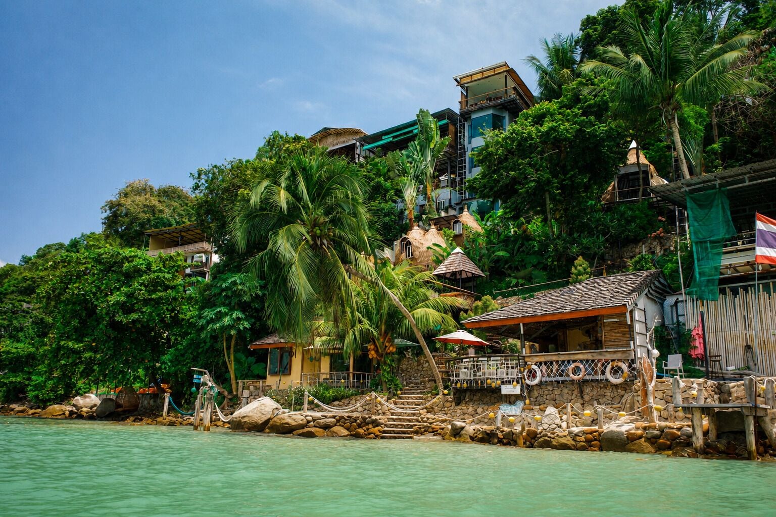 A coastal hotel with multiple buildings, including a prominent structure with a thatched roof and a smaller building with a tiled roof.
