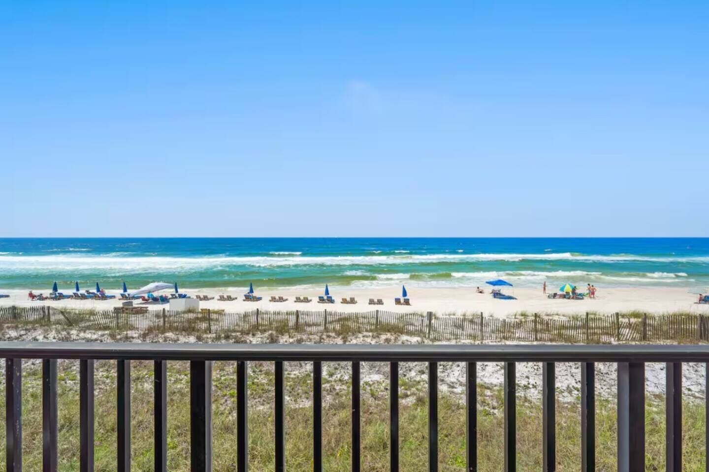 A beach with a wooden fence in the foreground, a clear blue sky, and the ocean in the background.