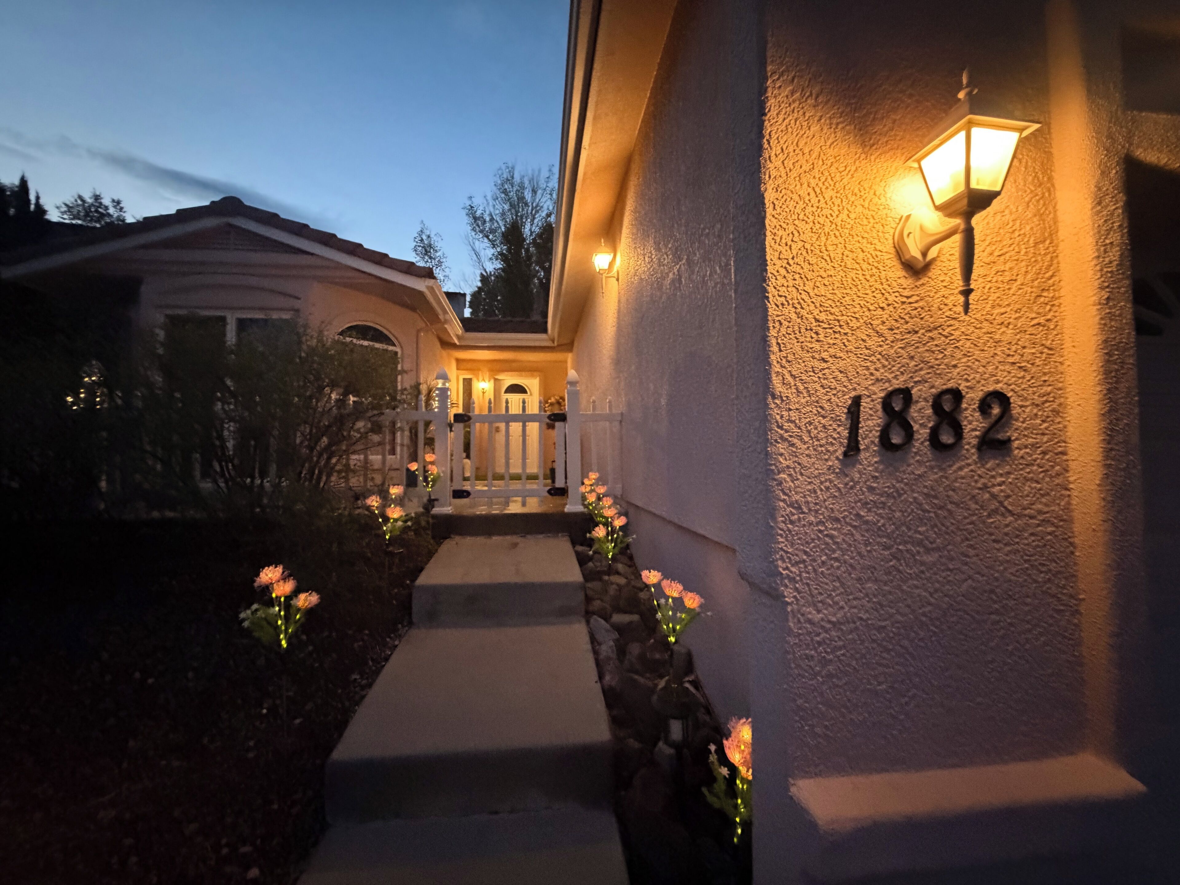A house with a well-lit entrance, a white picket fence, and a number '1882' on the wall.