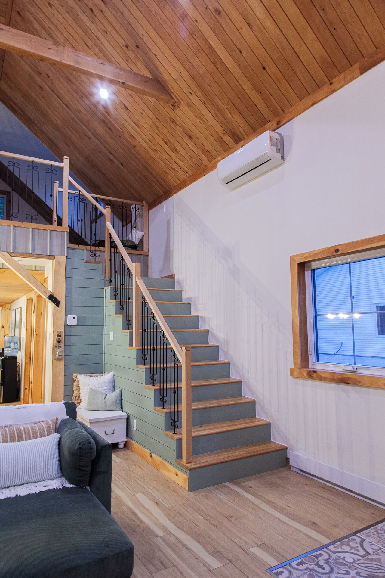 A staircase with wooden steps and metal railings, a white wall with a window, and a wooden ceiling.