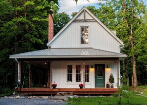 A white wooden house with a gabled roof, a brick chimney, and a front porch with potted plants.