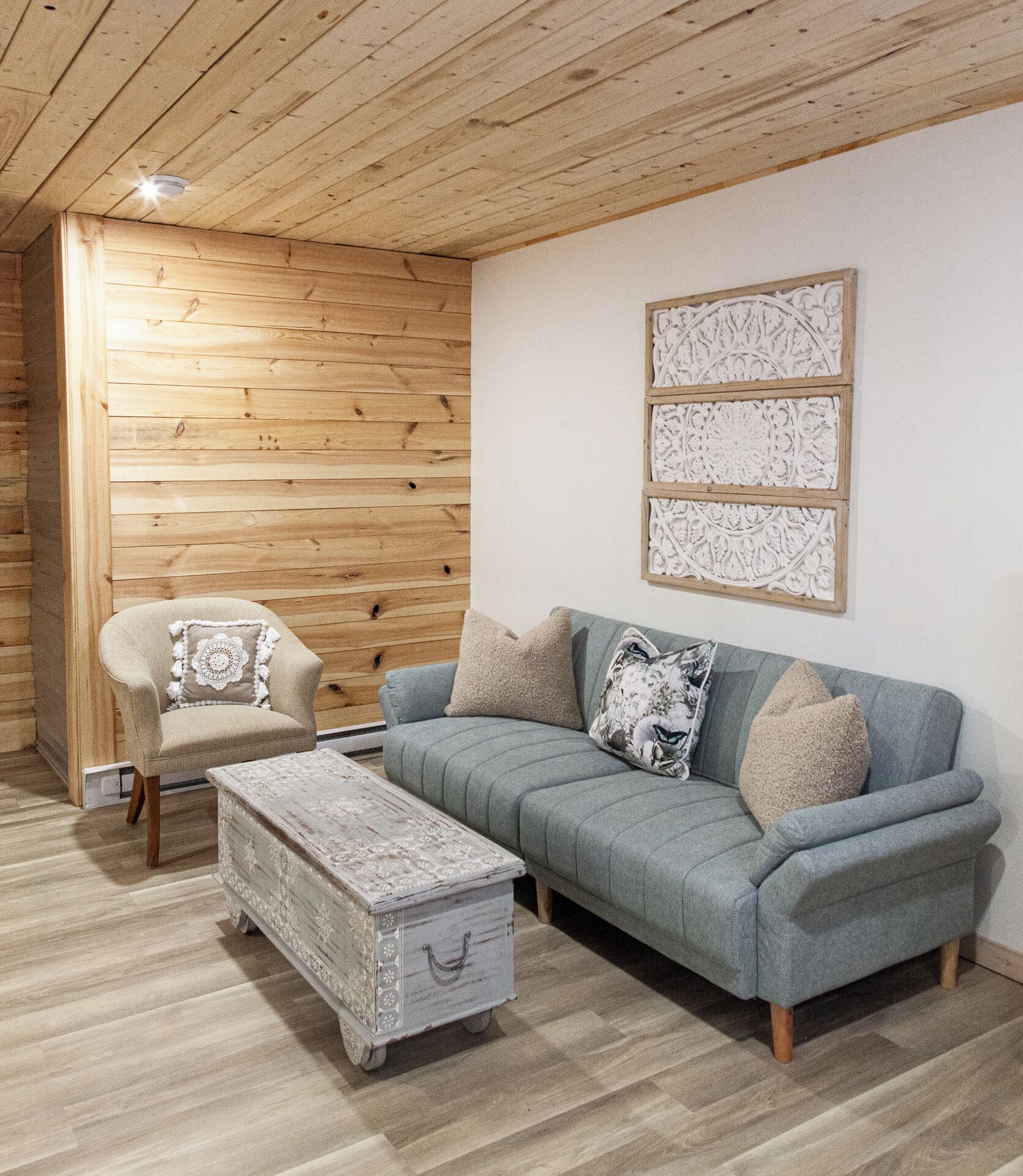 A cozy room with a wooden ceiling, a grey sofa with patterned cushions, a small armchair, and a decorative chest of drawers.
