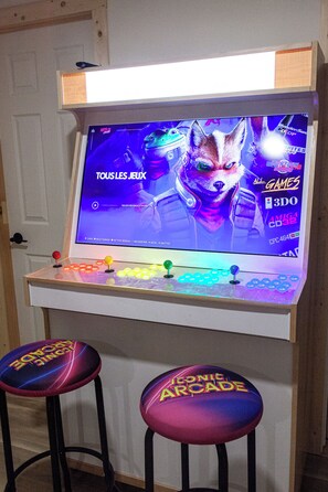 A gaming arcade with a large screen displaying a game, colorful arcade machines, and two stools with 'Iconic Arcade' branding.
