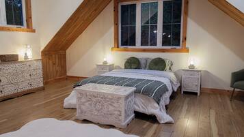 Una camera da letto con soffitto spiovente, un'ampia finestra, un letto con copriletto fantasia, due comodini con lampade e un settimino decorativo.