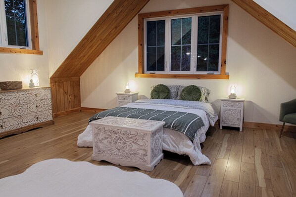 A bedroom with a sloped ceiling, a large window, a bed with a patterned bedspread, two bedside tables with lamps, and a decorative chest of drawers.