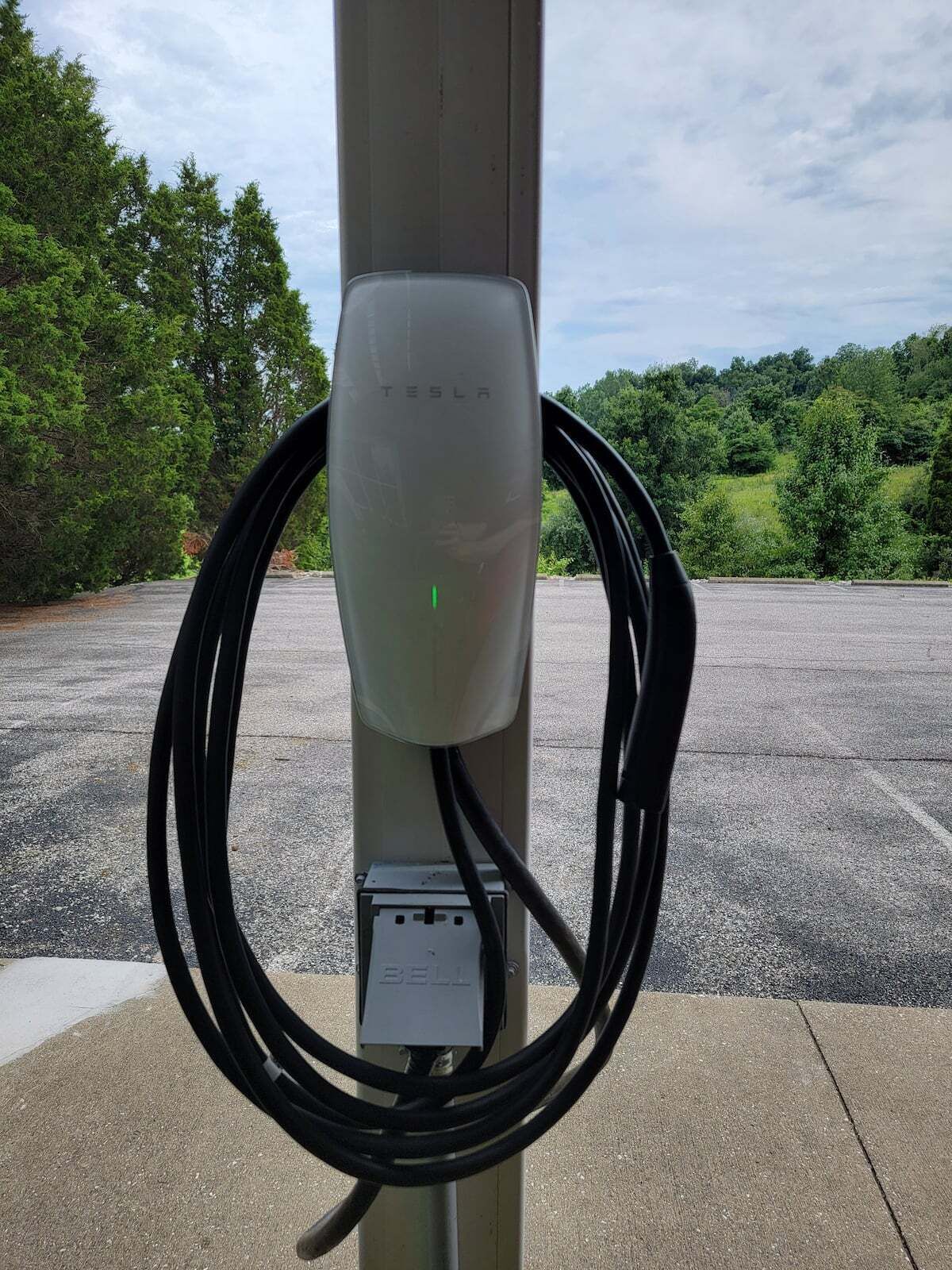 Eine Tesla-Ladestation mit einer darum gewickelten Ladekabel vor Bäumen und blauem Himmel.