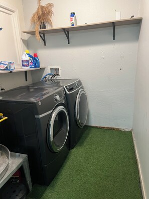 A laundry room with two front-loading washing machines, a shelf with cleaning supplies, and a small table with a basket.