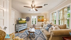 A living room with a sofa, a coffee table, a TV, a clock, and a ceiling fan. - The Coop & Roost | Hot Tub & Cowboy Pool! (Fredericksburg)
