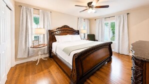 A bedroom with a wooden bed, a ceiling fan, and two windows with curtains. - The Coop & Roost | Hot Tub & Cowboy Pool! (Fredericksburg)