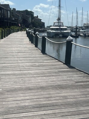 A wooden boardwalk with a fence on one side, leading to a marina with boats.